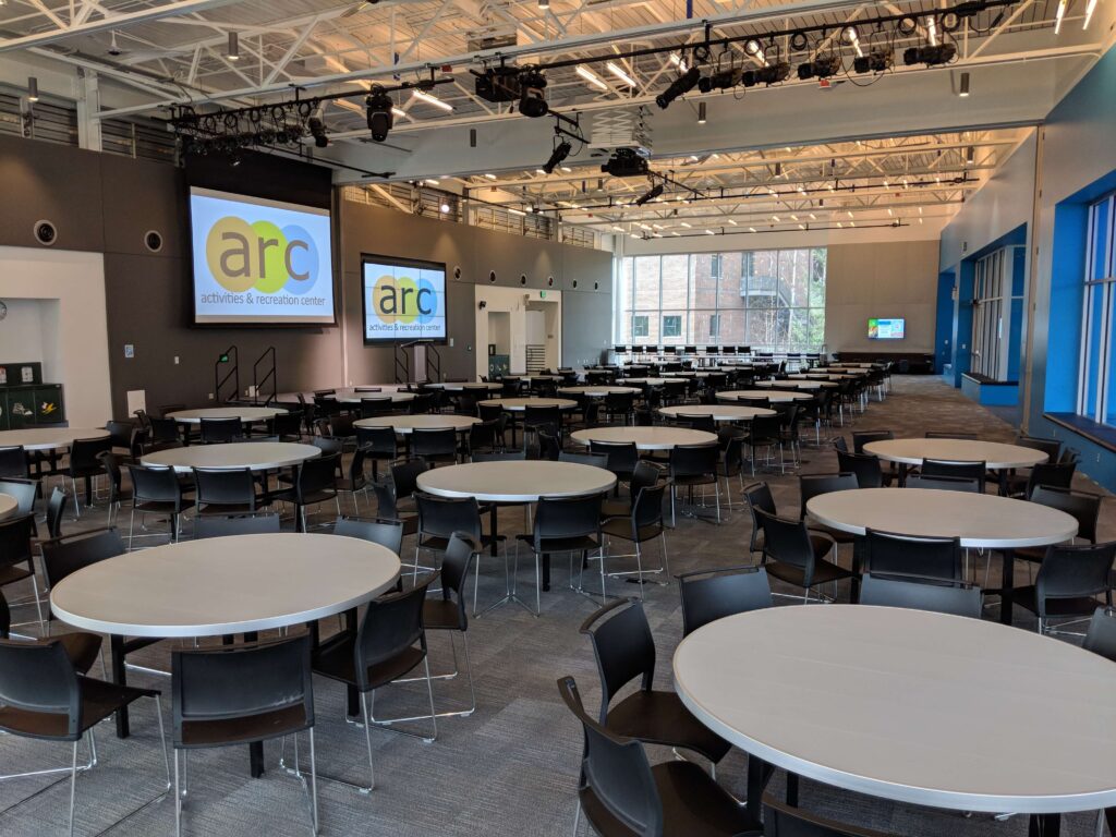 Image of room with circle tables with eight chairs each. Photo is taken from East side of room facing West.