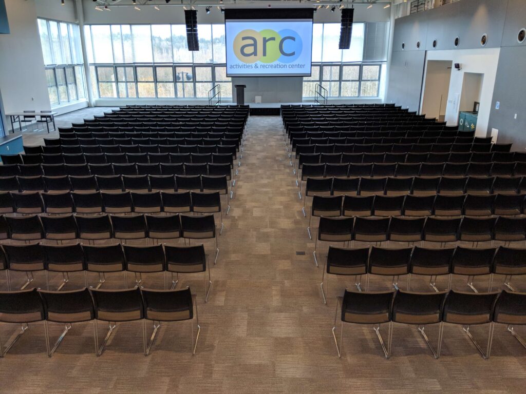 Image of room from West to East. Rows of chairs on right and left side with aisle in center. Projector screen and stage visible in center back of photo.