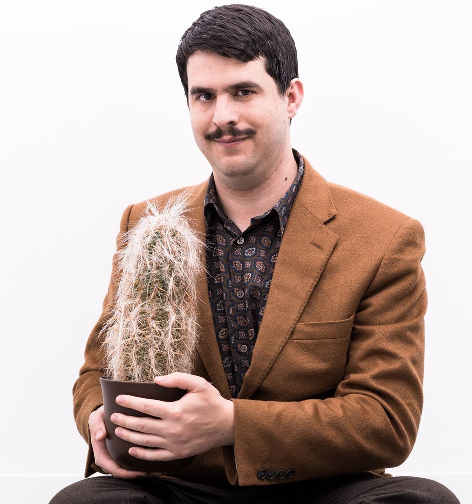Matthew Spencer poses in front of the camera with a potted cactus in hand. They are wearing a brown blazer and patterned button down shirt.