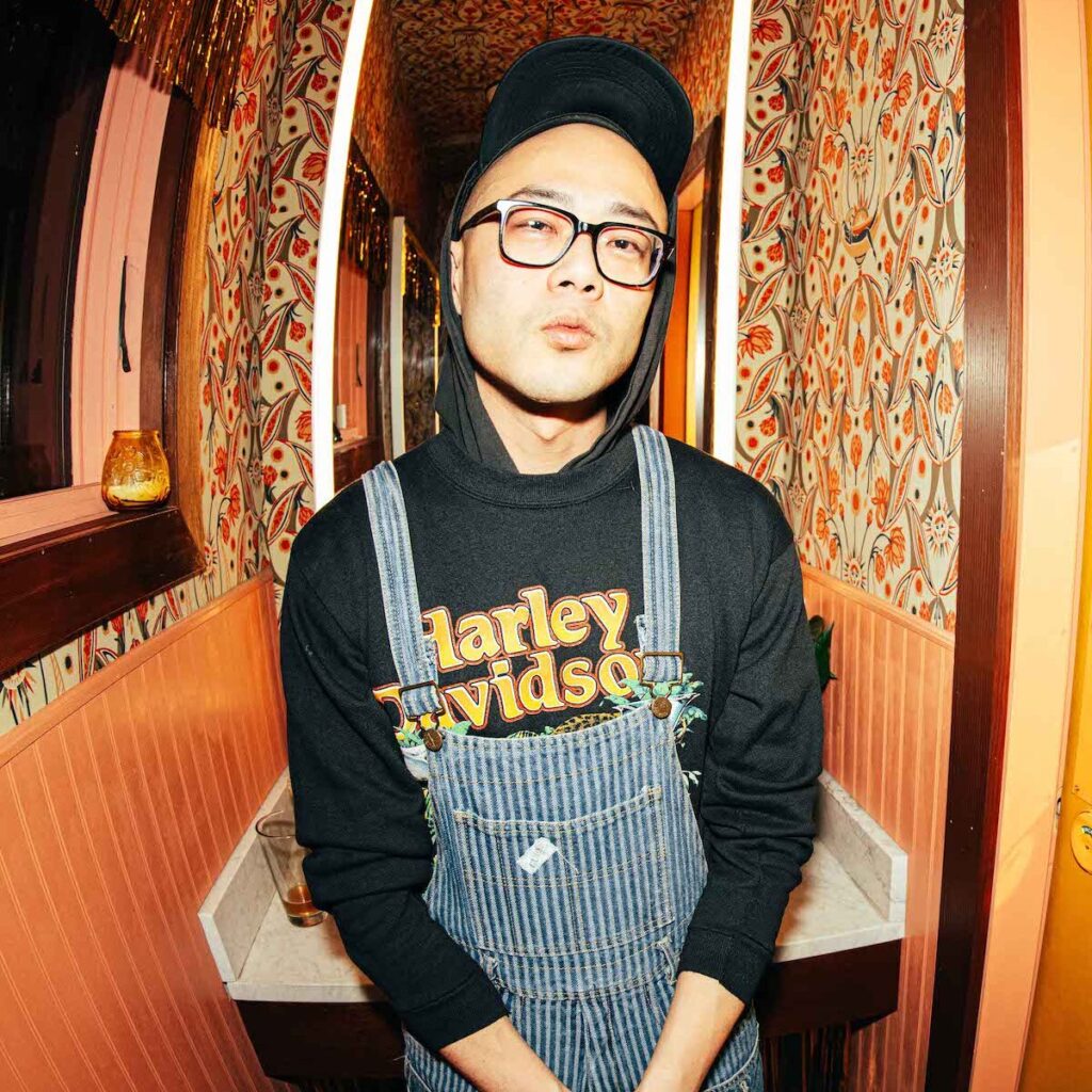 Richard Chiem wearing overalls, a baseball cap, glasses, and a Harley Davidson hoodie. He is posed in front of a hallway with patterned wallpaper.