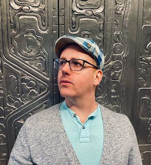 Evan Peterson in front of a metal glyph wall. Wearing a gray cardigan and a sky blue shirt and blue and white checkered hat