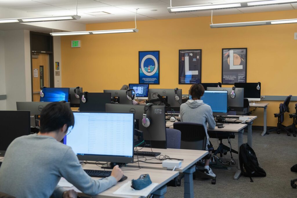 Open Learning Lab showing rows of adjustable computer desks (PCs and Macs), each computer with headsets, students working at some stations and the 3-poster artwork on the back wall spelling out O, L, L.