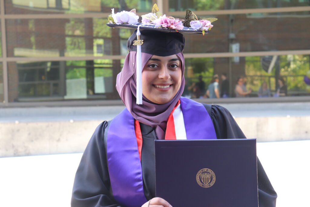 A person wearing a graduation cap and gown, holding up a degree holder.