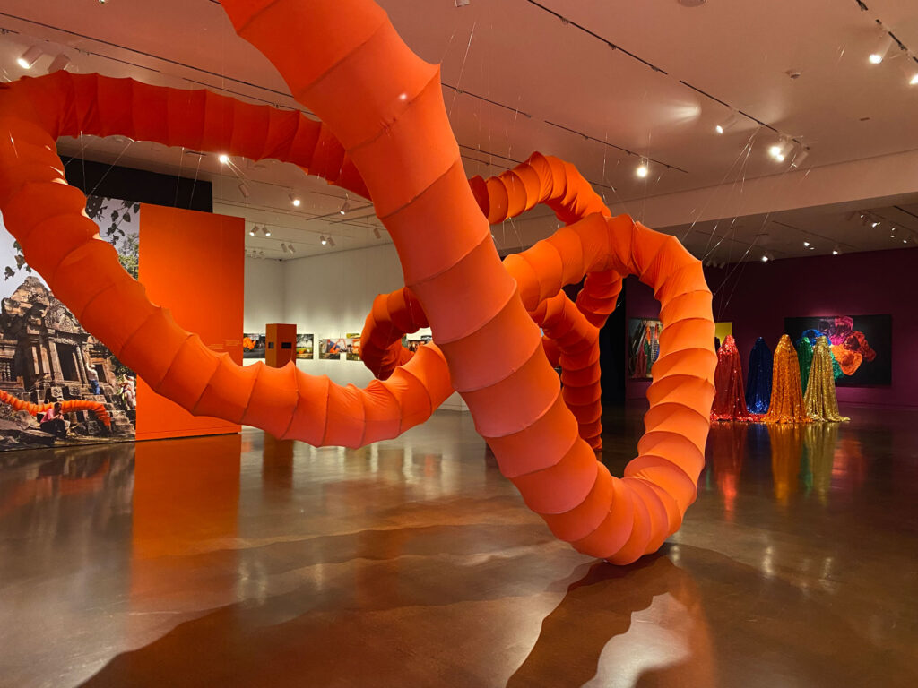 A long orange tube twists from floor to ceiling in a museum exhibition space.