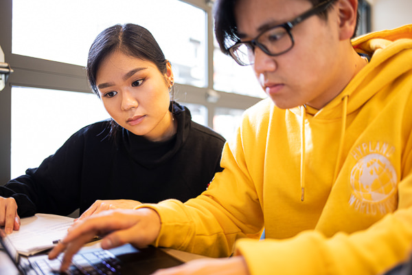 One male with dark hair, glasses and wearing a yellow hoodie and a dark haired woman with long sleeved black top looking at a computer screen.