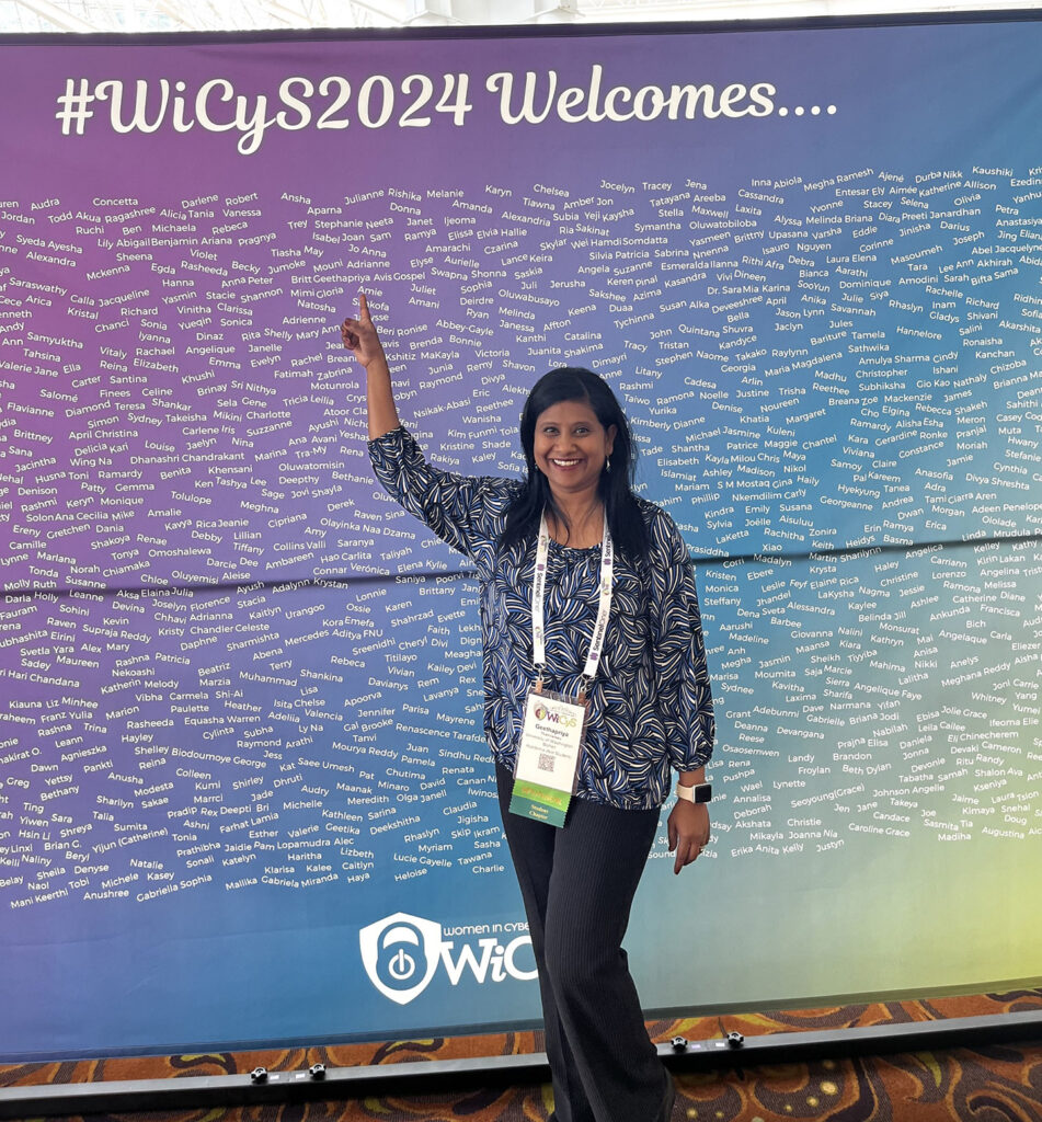 Geethapriya Thamilarasu pointing to her name on a large poster stating "#WiCyS2024 Welcomes...."