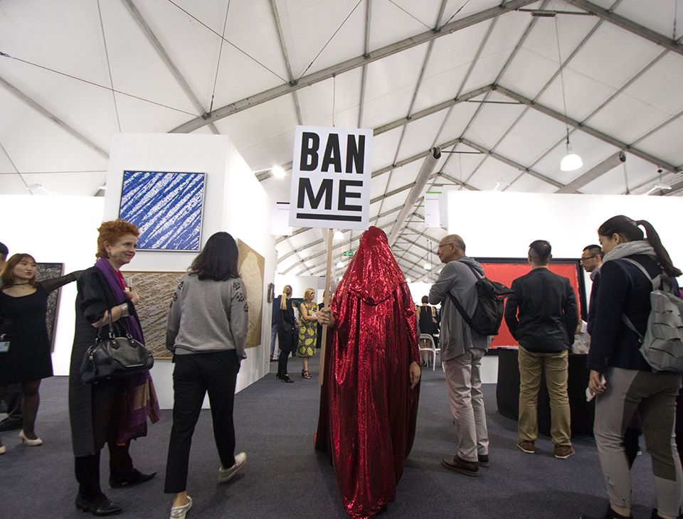 photo of the Red Chador holding a "Ban Me" sign while walking among people