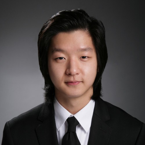 Tyler Choi in a black suit jacket, white button up, black tie with slicked back shoulder-length black hair.